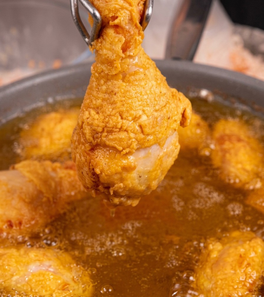 Una singola coscia di pollo fritta color dorato tenuta con pinze di metallo sopra una padella di olio bollente.