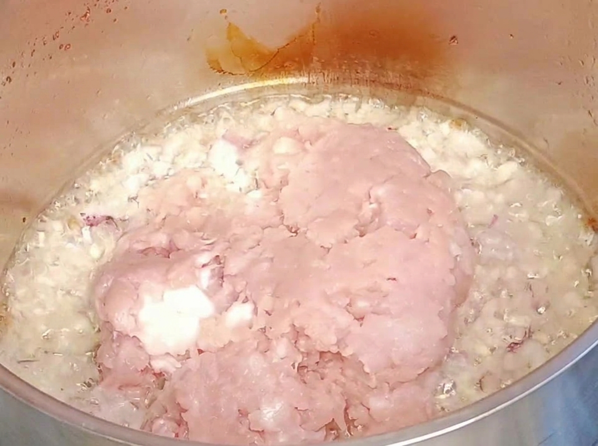 Carne macinata cruda aggiunta sopra cipolle e aglio rosolati in una pentola di metallo.