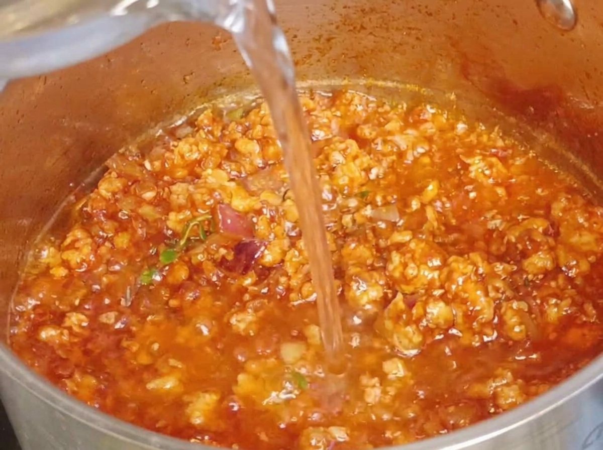 Acqua limpida versata in una pentola contenente il ricco ragù di carne.