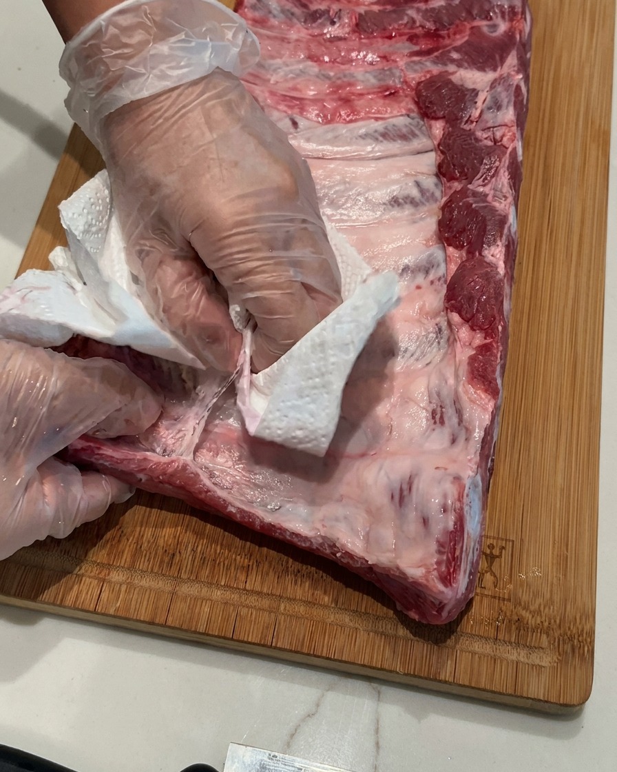 Mani con guanti in plastica che usano un tovagliolo di carta per rimuovere la sottile membrana da un rack di costine crude su un tagliere in legno.