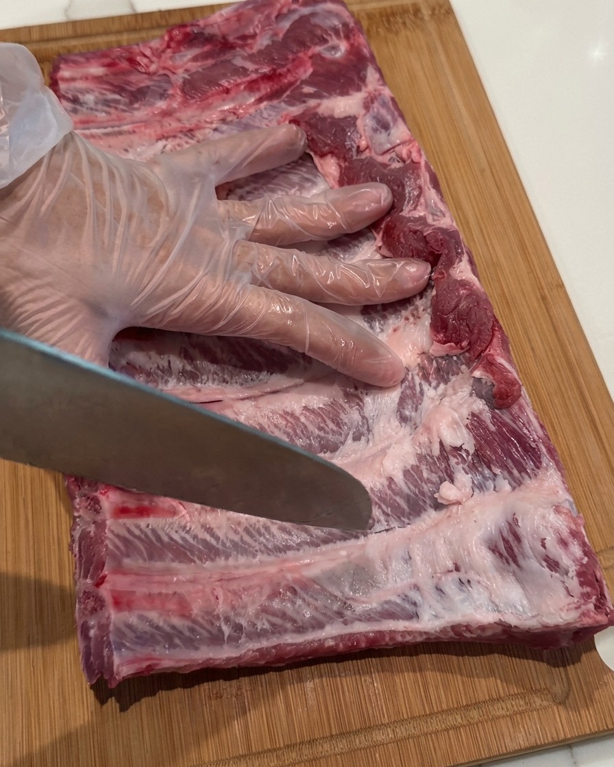 Una mano guantata che tiene un coltello da chef, usando la punta per praticare piccoli fori sulla superficie di un rack di costine crude.