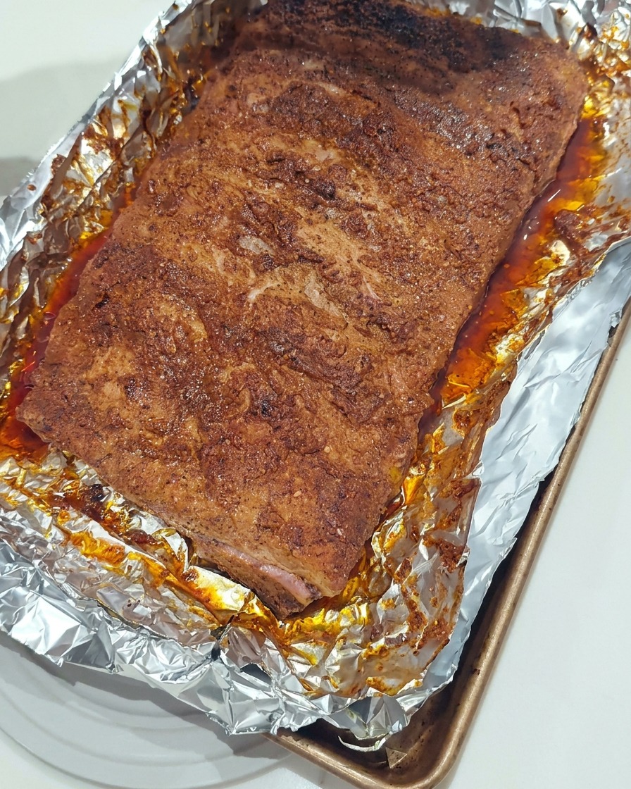 Un pennello in silicone che applica uno strato lucido di salsa BBQ densa sulla superficie delle costine arrostite in una teglia foderata con alluminio.