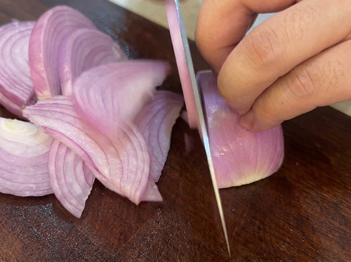 Un coltello che affetta una cipolla rossa in strisce su un tagliere di legno scuro.