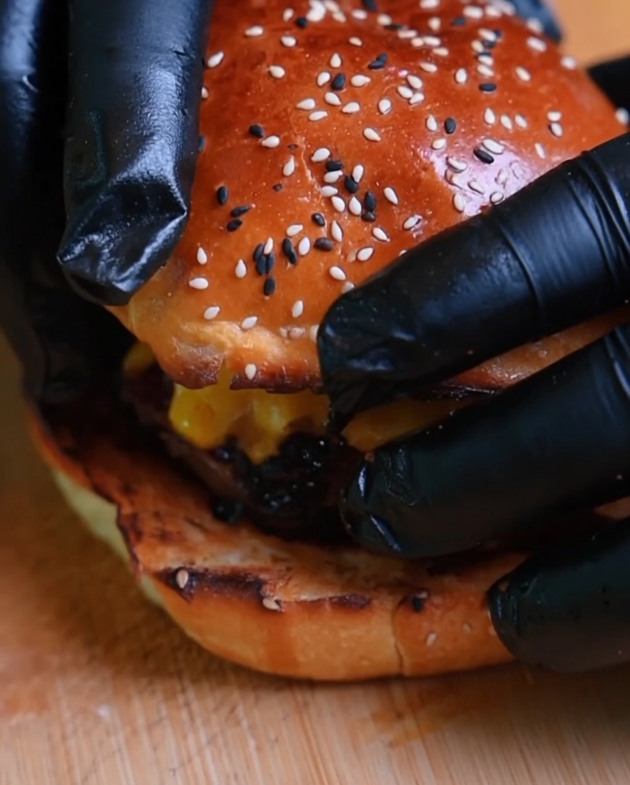 Un primo piano di un hamburger completato con il panino superiore posizionato sopra, tenuto da mani con guanti.