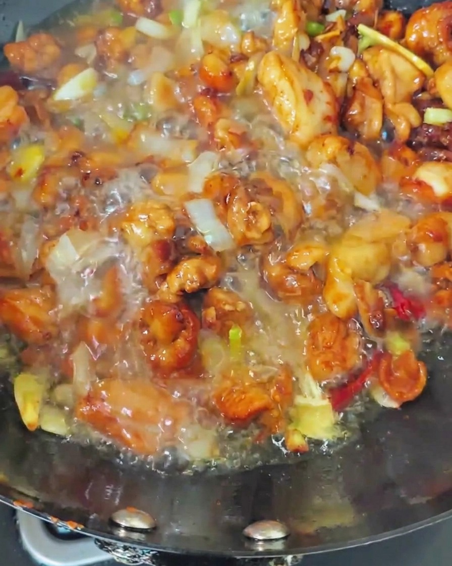 Acqua bollente versata nel wok contenente pollo rosolato e aromi, creando un brodo gorgogliante.