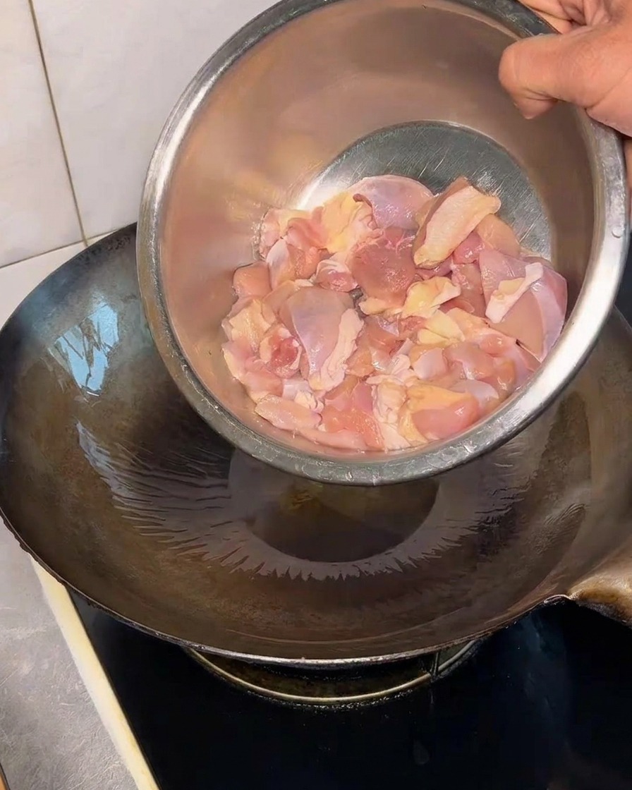 Pezzi di pollo crudo versati da una ciotola di metallo in un wok rovente oliato.