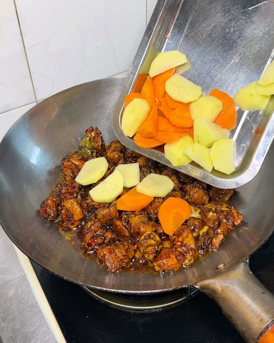 Pezzi di patate e carote versati da un vassoio nel wok con pollo e salsa scura.