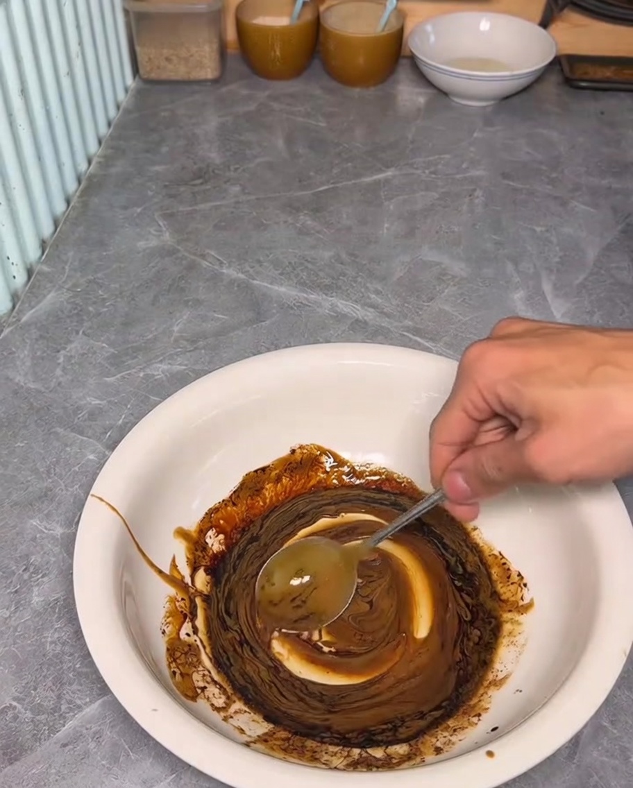 Una mano usa un cucchiaio per mescolare salsa di sesamo e soia scura in una ciotola bianca.
