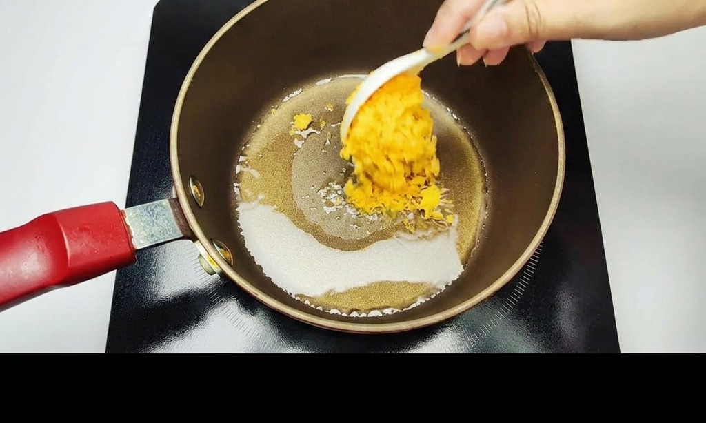 Una mano che utilizza un cucchiaio bianco per mescolare i tuorli duovo salati dorati e schiacciati in una pentola antiaderente scura.