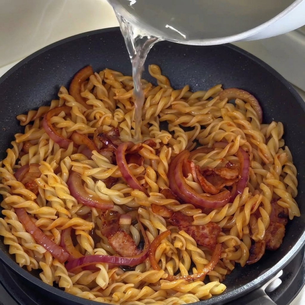 Acqua di cottura della pasta versata da un pentolino nella padella con i fusilli.