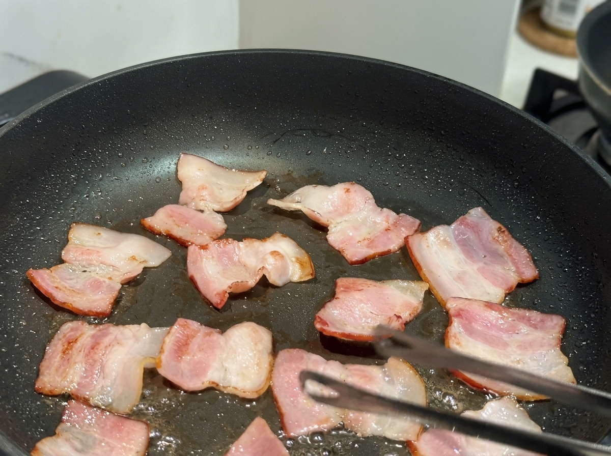 Piccoli pezzi piatti di pancetta in cottura in una padella antiaderente nera, con una pinza visibile.