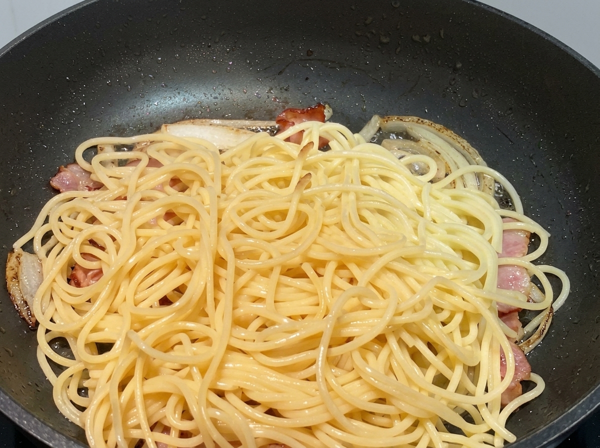 Spaghetti cotti impilati sopra cipolle rosolate e pezzi di pancetta in una padella nera.