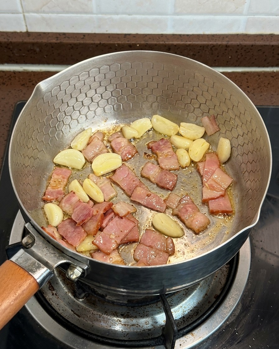 Funghi affettati, pezzi di pancetta e spicchi daglio che cuociono insieme in padella.