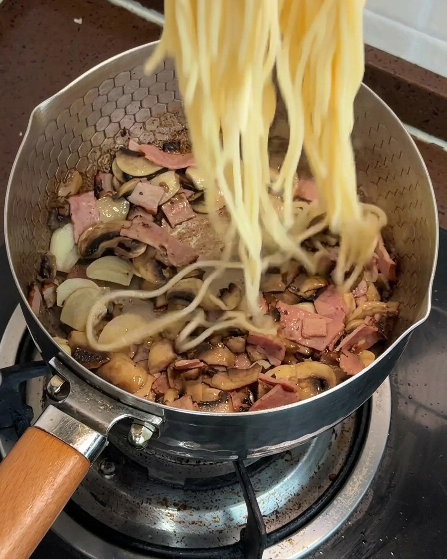 Spaghetti cotti che vengono aggiunti in una padella piena di funghi e pancetta saltati.