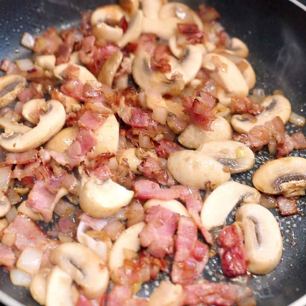Pasta lunga aggiunta in una padella piena di pancetta dorata, funghi saltati e cipolle.