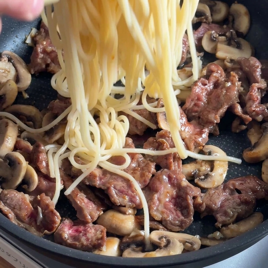 Pasta aggiunta alla padella con manzo e funghi, pronta per essere condita.