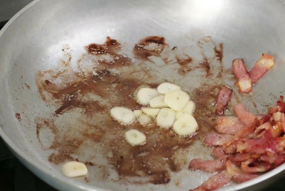 Aglio a fette e funghi che rosolano in padella insieme alla pancetta croccante.