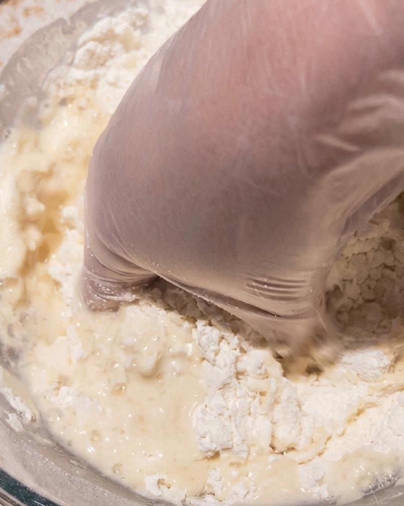 Una mano guantata che mescola limpasto appiccicoso e umido in una grande ciotola di vetro.