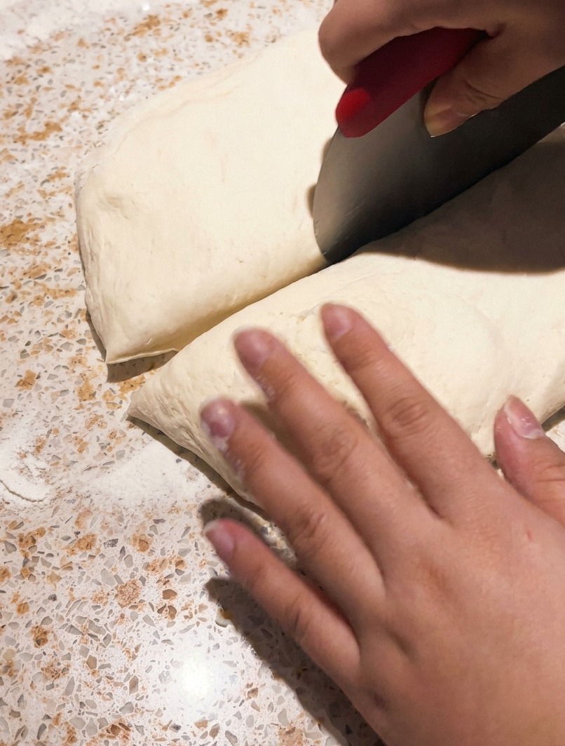 Un raschietto da banco in metallo che taglia un pezzo di impasto per pizza fermentato su un piano di lavoro.