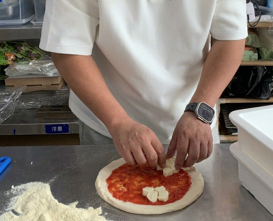 Mani che posizionano cubetti di mozzarella fresca su una base di pizza con salsa.