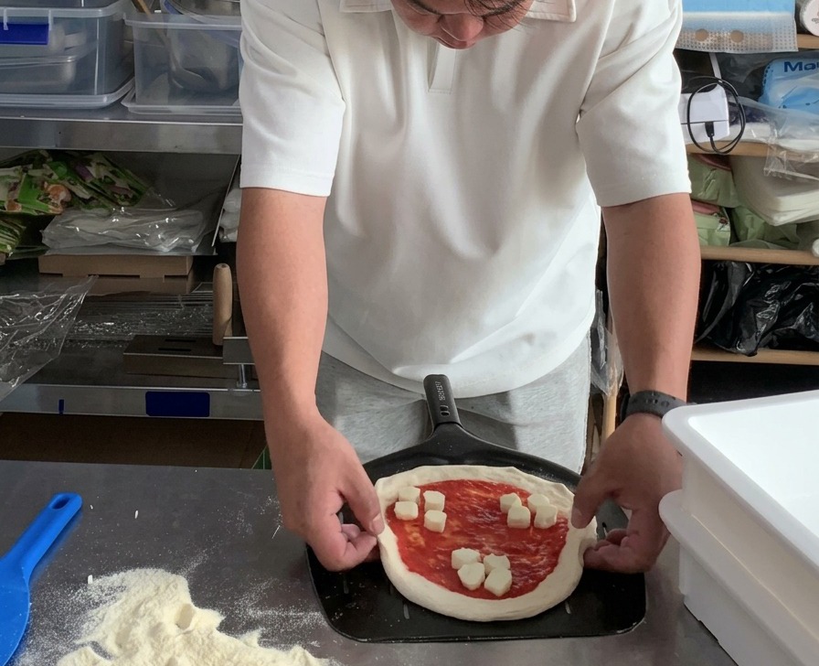 Mani che sistemano i bordi di una pizza Margherita cruda su una pala forata in metallo.