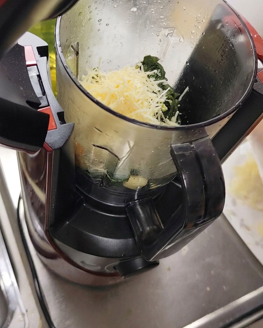 Ingredienti come formaggio grattugiato, aglio e pinoli inseriti in un robot da cucina insieme alle foglie di basilico.