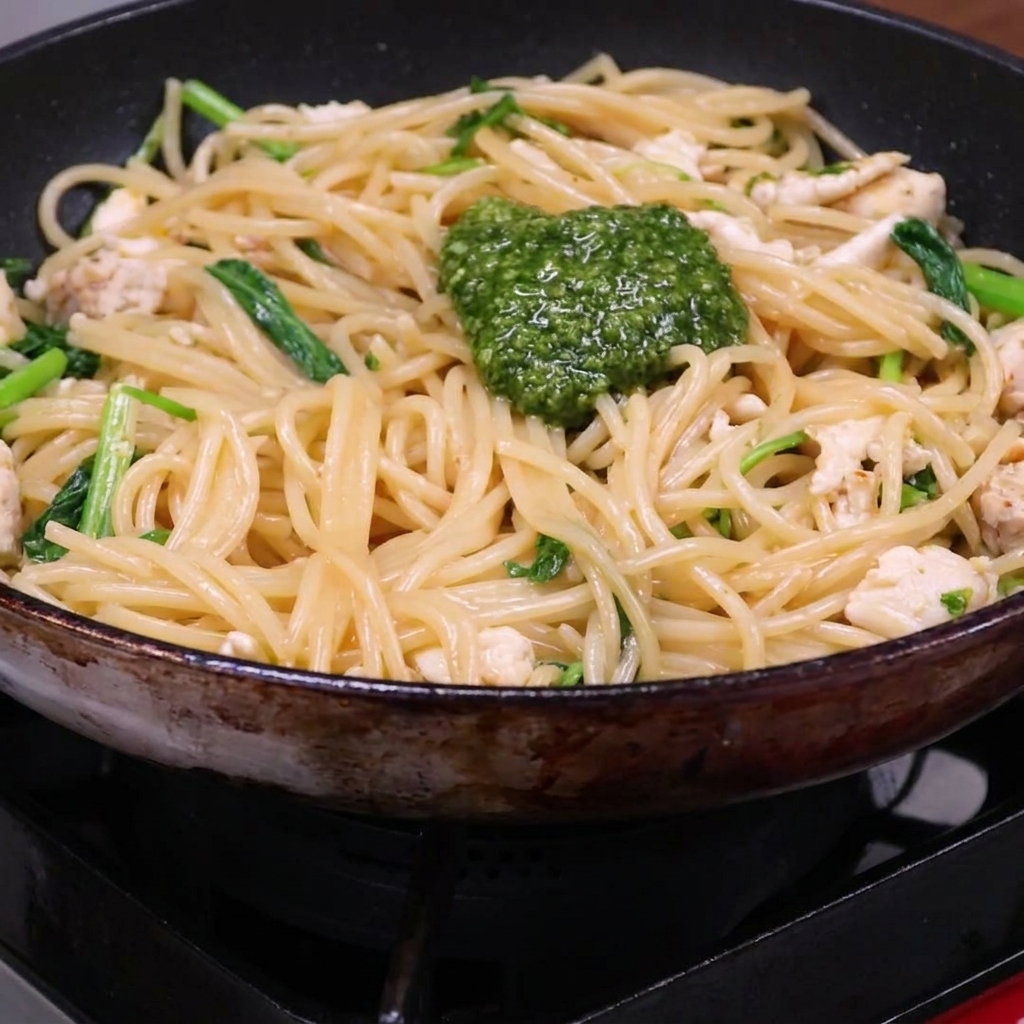 Un cucchiaio di pesto verde brillante aggiunto a un mix di spaghetti cotti, pollo e rucola in padella.