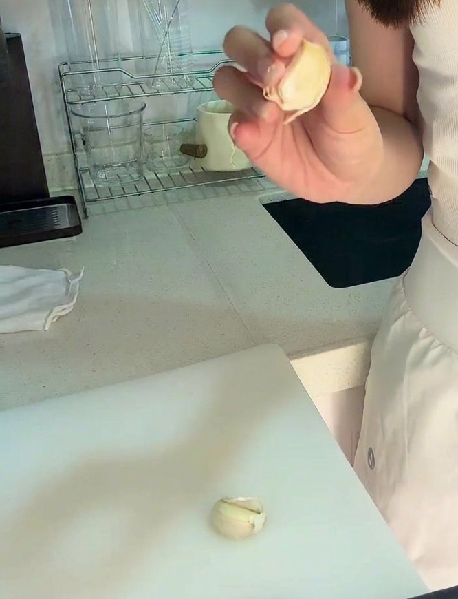 Una mano che tiene uno spicchio daglio sbucciato su un tagliere bianco con una piccola quantità di aglio tritato visibile.