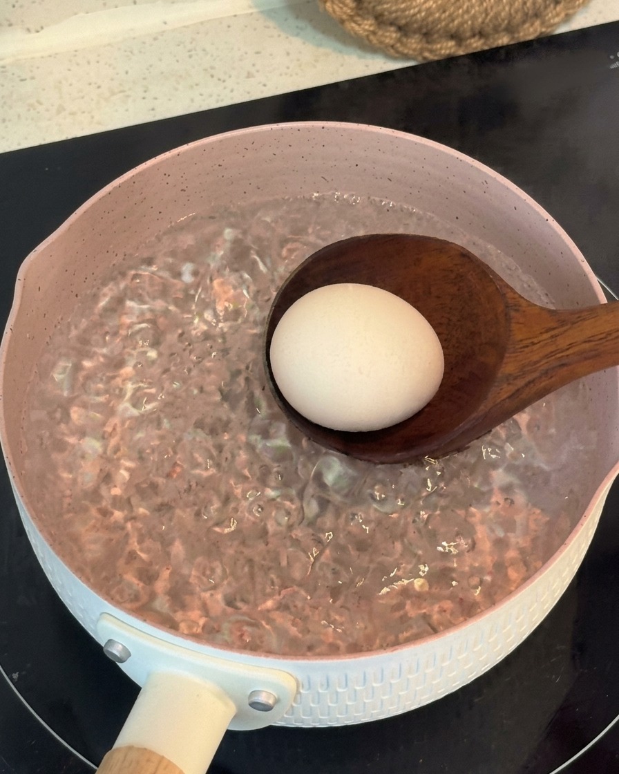 Un mestolo di legno che immerge delicatamente un uovo intero in una pentola rosa di acqua in piena ebollizione.