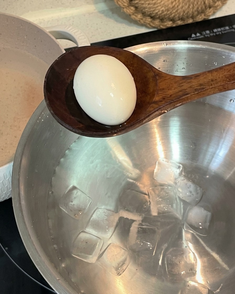 Un cucchiaio di legno che trasferisce un uovo sodo in una ciotola di acciaio inossidabile riempita con acqua fredda e cubetti di ghiaccio.