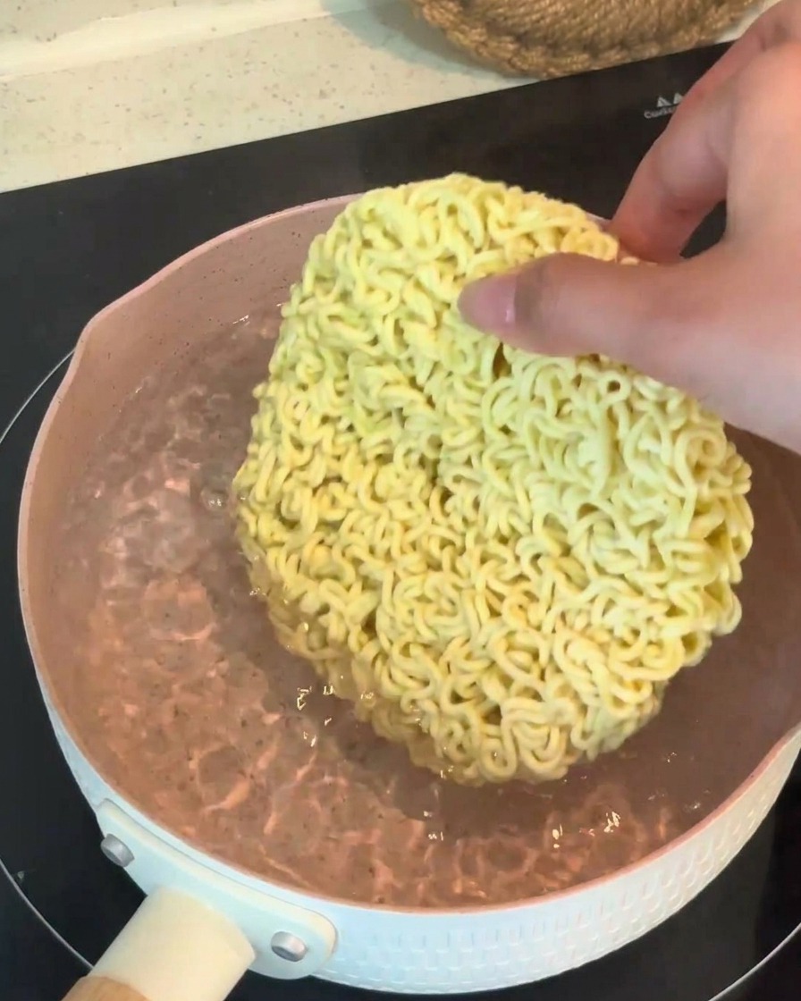 Una mano che mette un panetto di noodles ramen istantanei secchi in una pentola di acqua bollente.