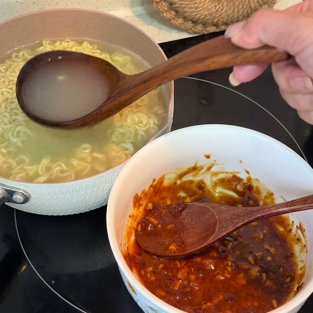 Acqua calda dei noodles versata da un mestolo di legno in una ciotola contenente una pasta di condimento mescolata.