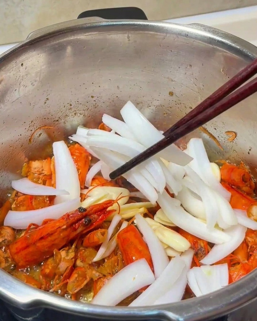 Un primo piano di fette di aglio e strisce di cipolla mescolate ai carapaci di gambero fritti in una pentola di metallo.
