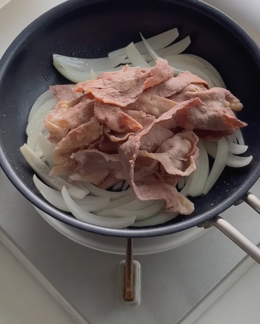 Fette di manzo sbollentate adagiate sopra le cipolle saltate in una padella nera.