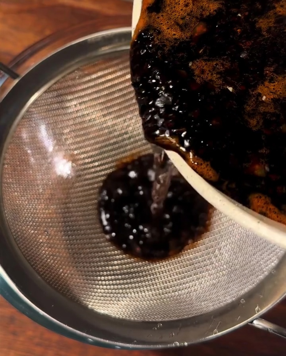 Salsa marrone scura versata attraverso un colino in una ciotola di vetro.
