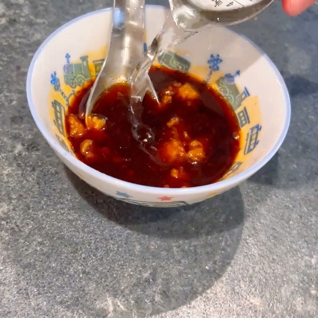 Acqua versata in una ciotola contenente salsa di soia scura e grumi di miso, con un cucchiaio che mescola il composto.