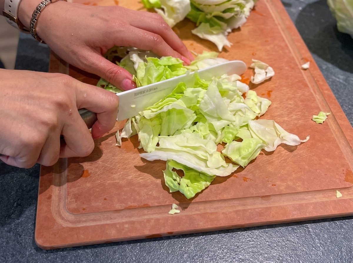 Mani che usano un coltello in ceramica bianco per tritare cavolo verde fresco su un tagliere di legno.