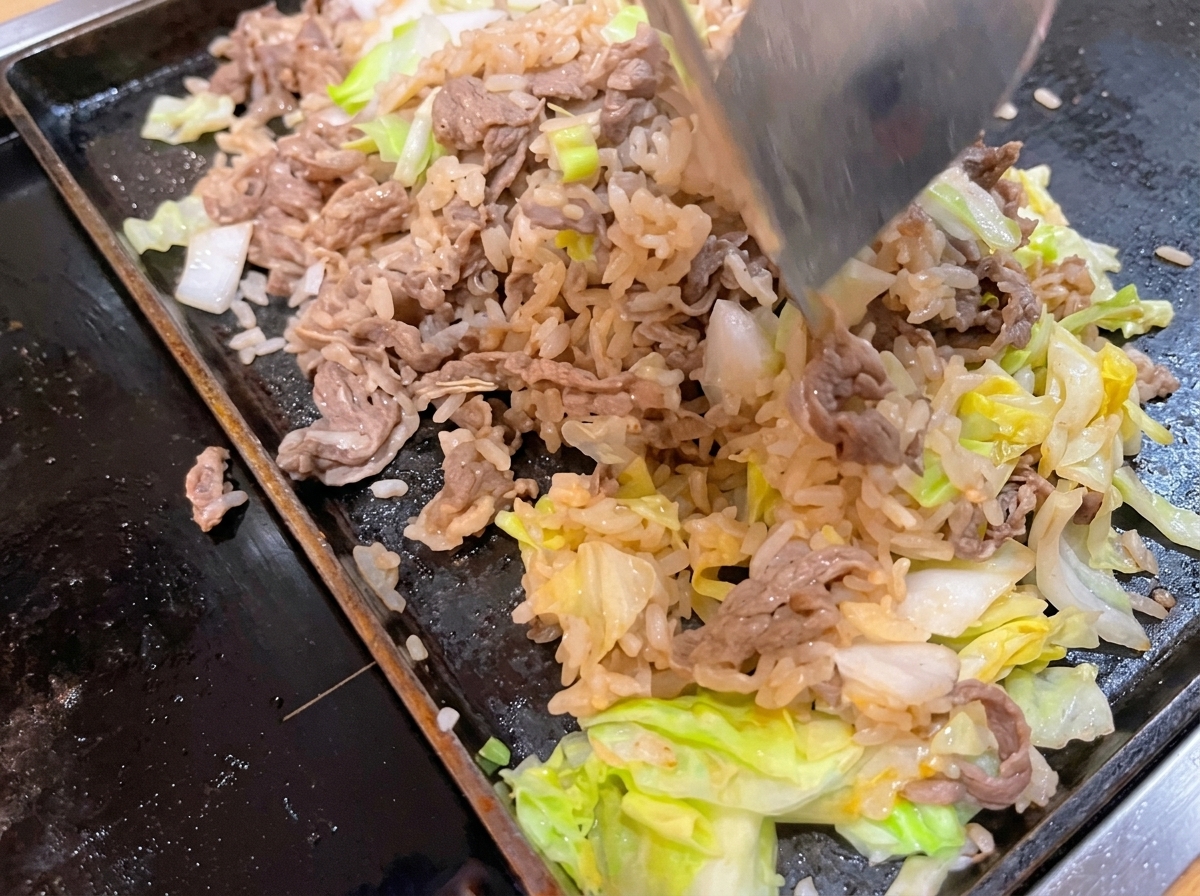 Una spatola di metallo che gira e mescola manzo, riso bianco e cavolo verde insieme su una padella piatta calda.