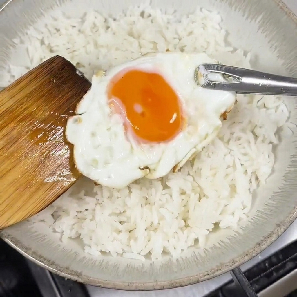 Una spatola di legno adagia un uovo fritto con tuorlo liquido su una ciotola di riso bianco.