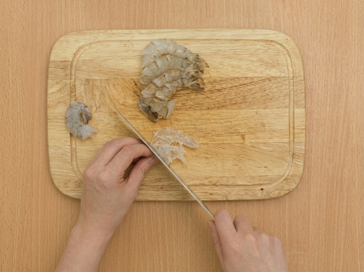 Primo piano di un coltello che pratica tagli superficiali lungo il ventre di un gambero crudo su un tagliere di legno.