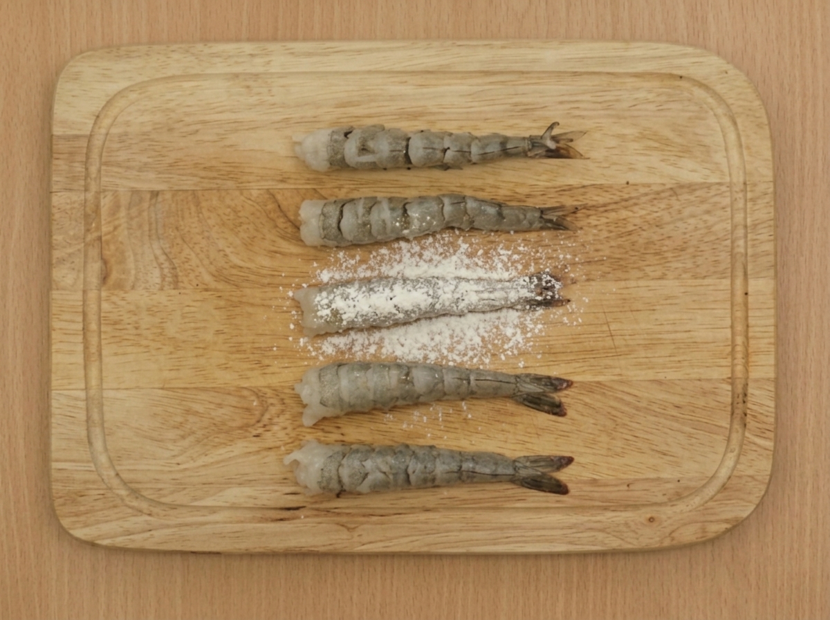 Un setaccio che spolvera farina debole su diversi gamberi preparati e disposti su un tagliere di legno.