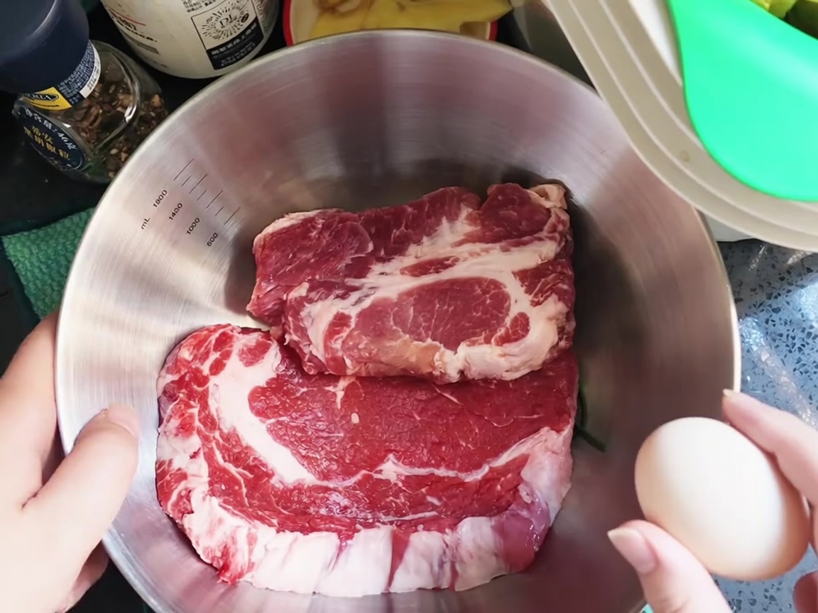 Un uovo crudo che viene rotto sopra delle braciole di maiale fresche in una ciotola di acciaio inox.