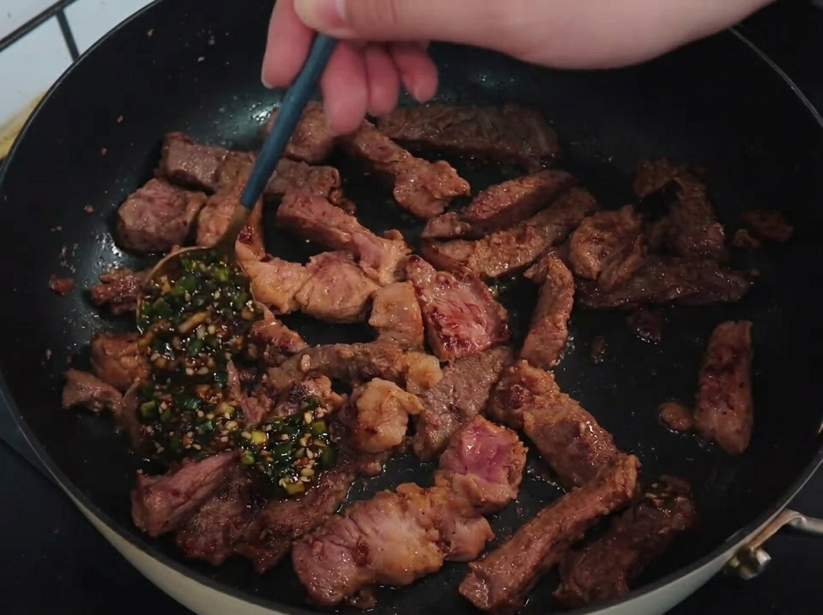 Un cucchiaio che versa una vibrante salsa verde di cipollotto e aglio su strisce di braciola di maiale rosolate in una padella calda.