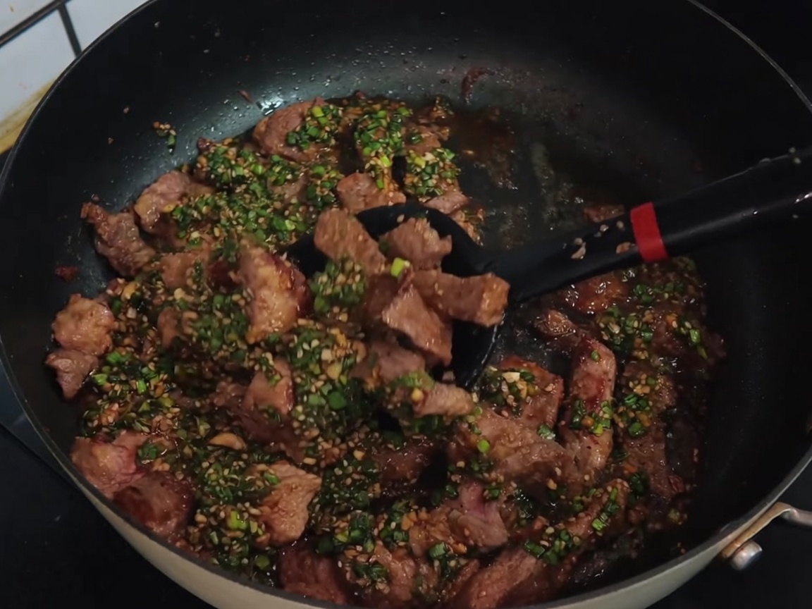 Una spatola nera che mescola strisce di braciola di maiale con una salsa verde al cipollotto in una padella antiaderente.
