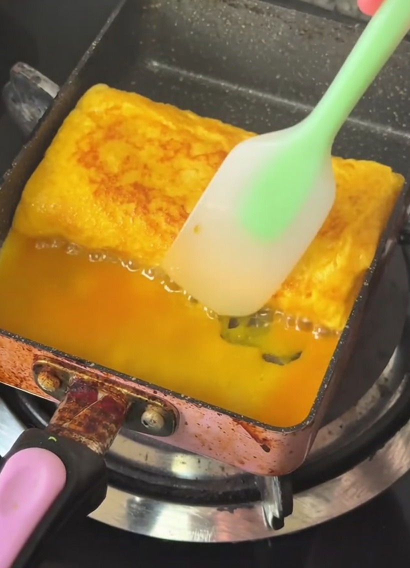Una padella per tamagoyaki con un rotolo duovo dorato a strati che viene ripiegato con una spatola in silicone.