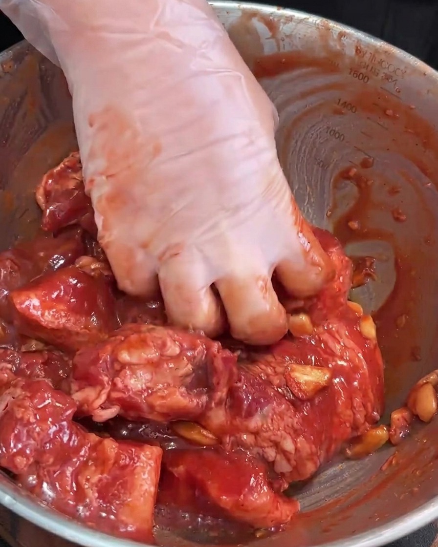 Mani con guanti che massaggiano la carne cruda e la marinata in una ciotola di acciaio.