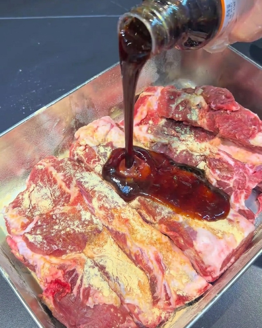 Salsa scura versata da una bottiglia su costine condite in una teglia.