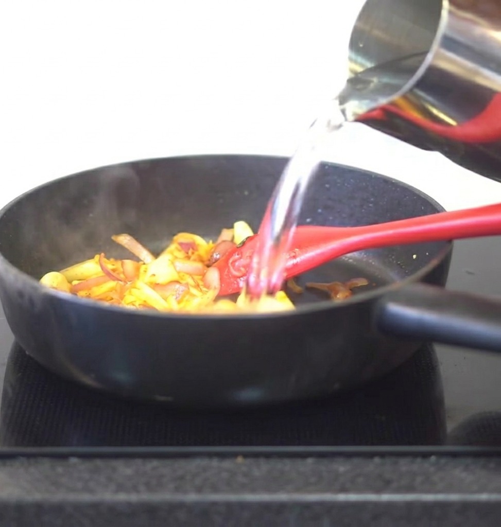 Acqua versata da una brocca di metallo in una padella contenente kimchi saltato e aromi.