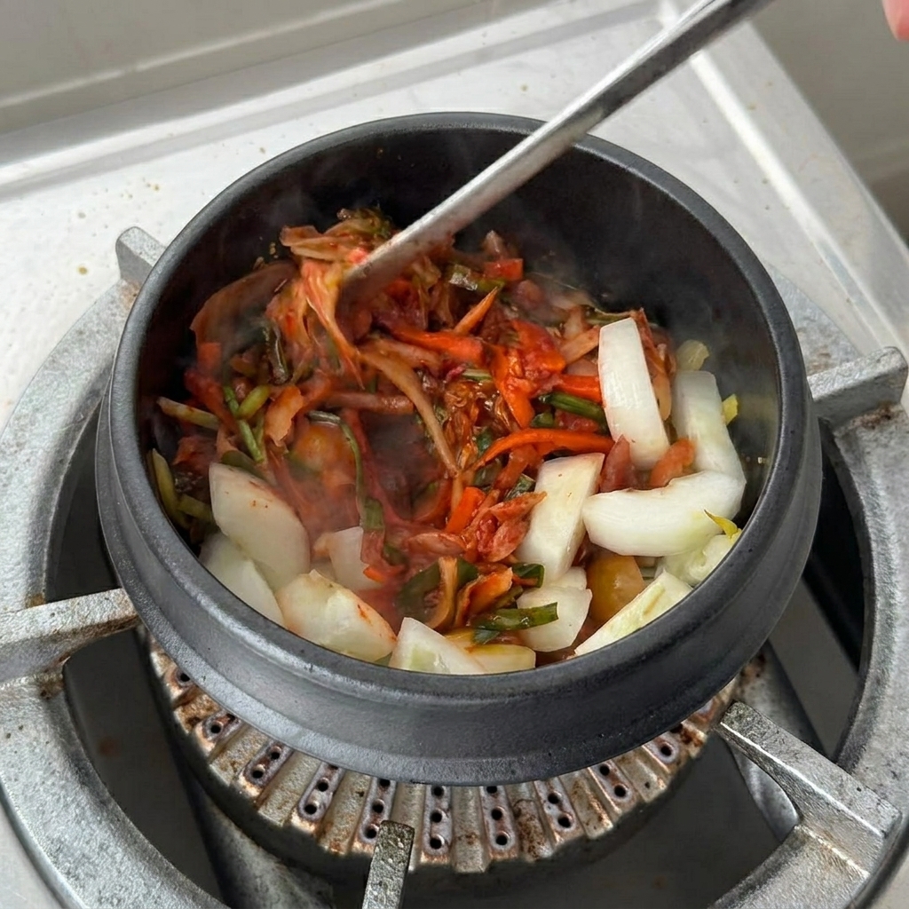 Una tazza grande di base di zuppa di kimchi versata nella pentola di terracotta sopra il kimchi e le cipolle soffritte.