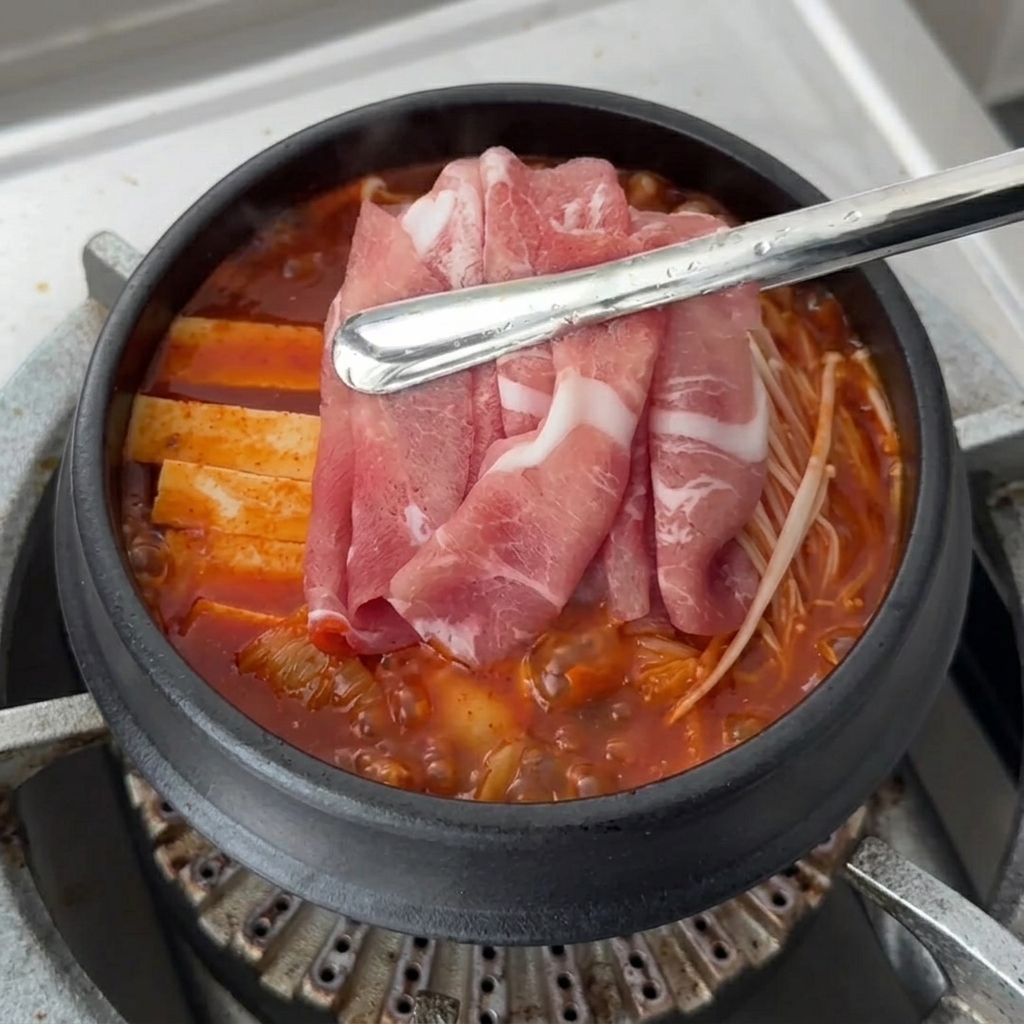 Fette di maiale crudo fresco disposte sopra gli ingredienti del Kimchi Jjigae in ebollizione.